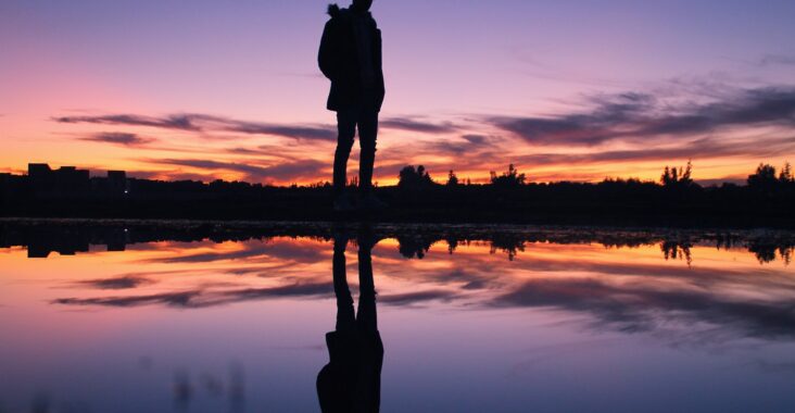 Image shows a male silhouette reflected in a body of water at dusk with a colourful sunset in the background